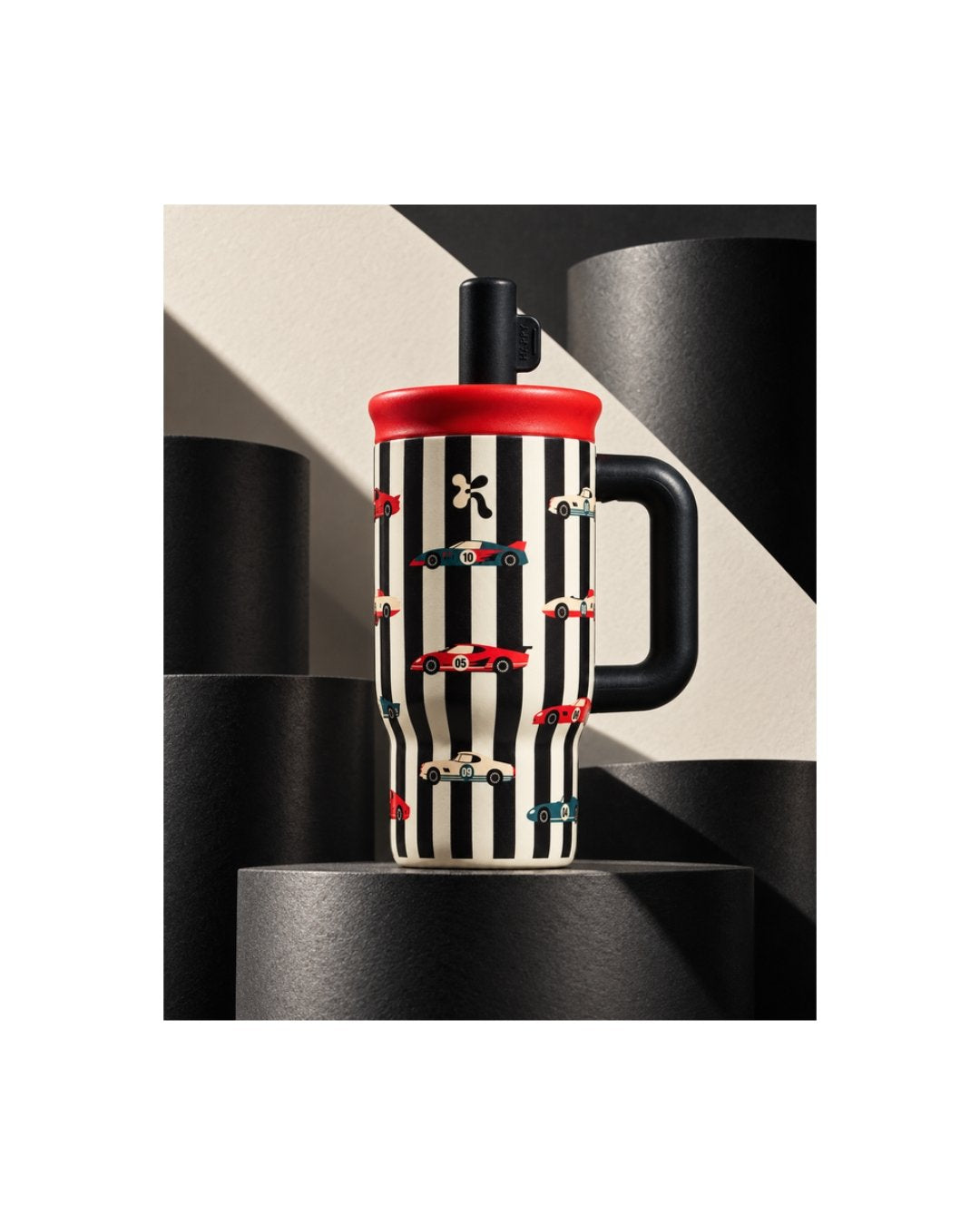 Tumbler Kara Bottles bergaris hitam putih bermotif mobil balap merah, tutup merah, sedotan dan gagang hitam, di atas podium hitam dengan latar krem