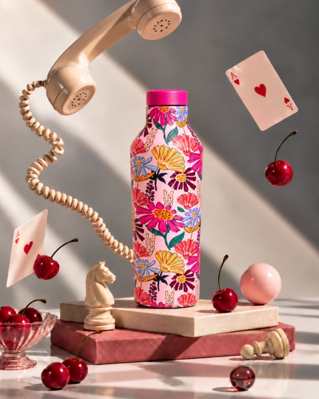 Tumbler stainless Kara Bottles bermotif bunga warna-warni dengan tutup pink, berdiri di atas balok di antara buah ceri, kartu remi, dan telepon vintage